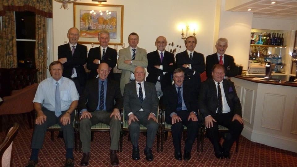 A group of people standing and sitting for a group photo. People are wearing suits. They are indoors and there is a painting on the wall behind them.