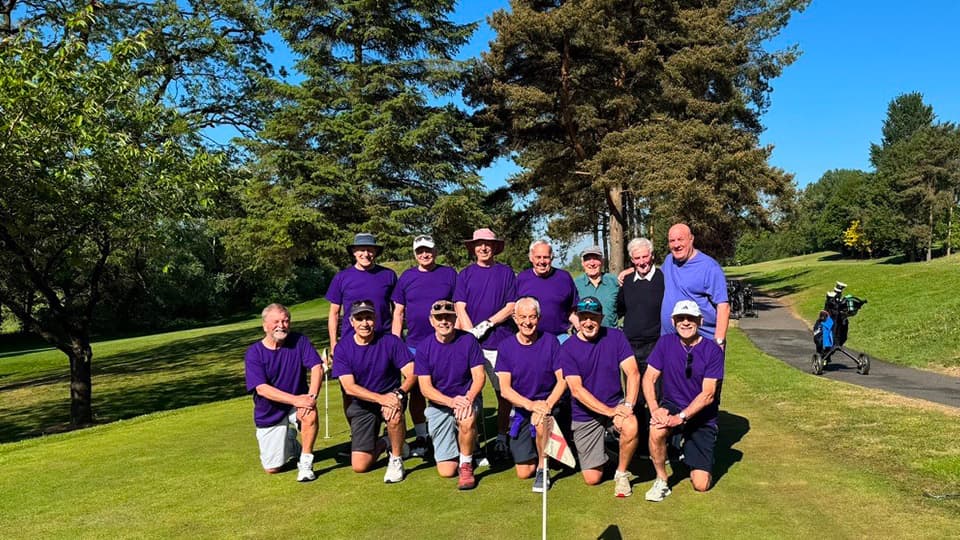 A group of people standing and crouching for a group photo. Most of the people are wearing African Violet t-shirts. They have golf clubs. There is grass and trees.