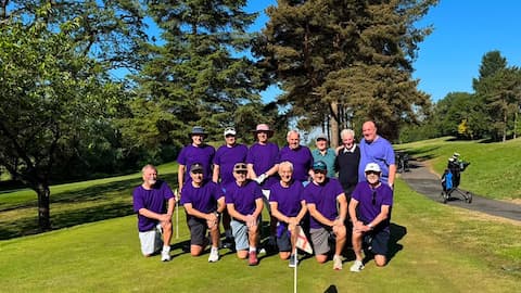 A group of people standing and crouching for a group photo. Most of the people are wearing African Violet t-shirts. They have golf clubs. There is grass and trees.