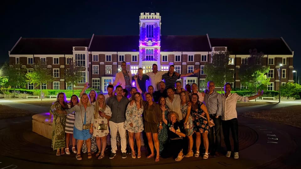 A large group of people standing in front of the Hazlerigg Building on campus. Many are on the ground, whilst some appear above them, standing on the wall of the fountain. It is night time.