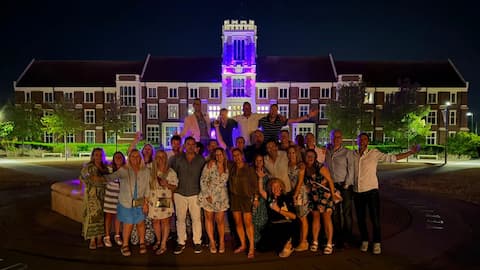 A large group of people standing in front of the Hazlerigg Building on campus. Many are on the ground, whilst some appear above them, standing on the wall of the fountain. It is night time.