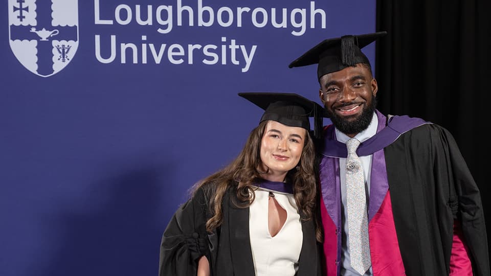 Libby and Jaivani wearing graduation attire. They stand in front of an African Violet backdrop that has a Loughborough University logo on.