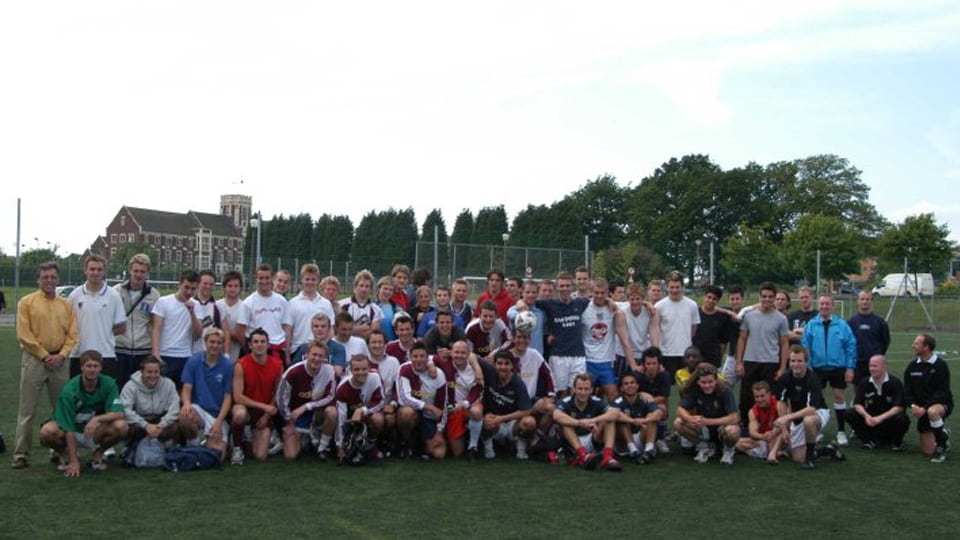 A large group of people crouched and standing together on grass. They are wearing sports clothes. Behind them is the University's Hazlerigg Building and some trees.