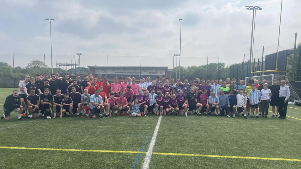 A large group of people crouched and standing together on grass. They are wearing sports clothes. Behind them is the University's football Stadium.