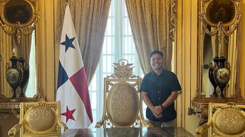 Mario standing beside an ornate desk in a grand room with gold decorative furnishings, large windows, and a Panamanian flag displayed behind the desk.
