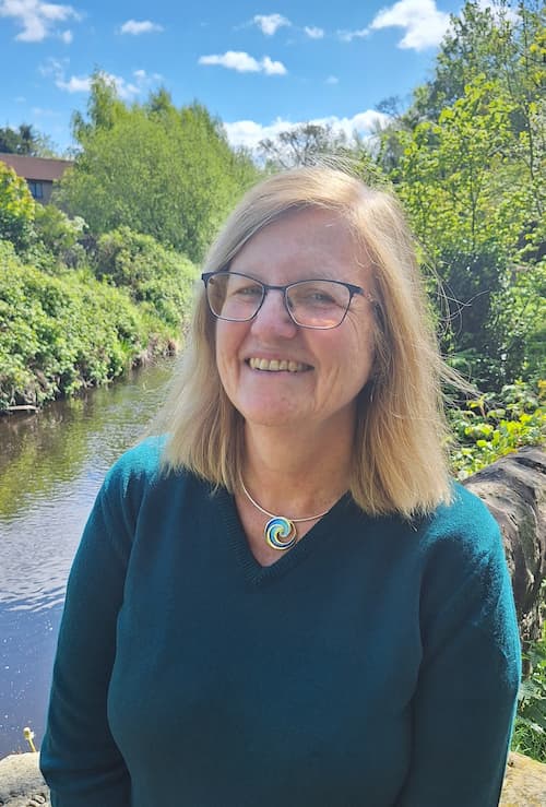 Sue Bell, smiling. She is outside. There is blue sky and trees behind her.