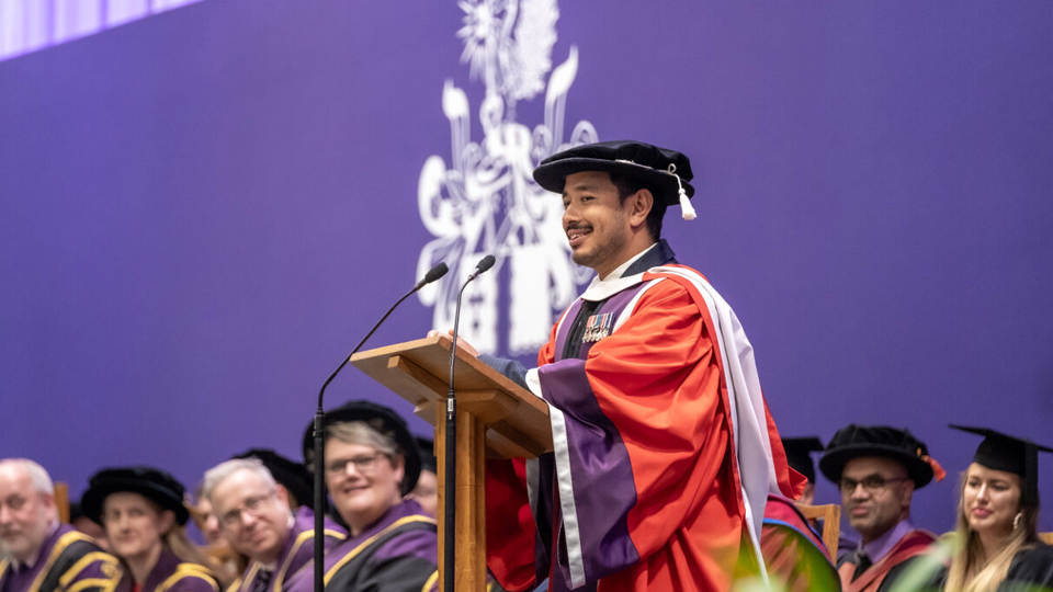 Nirmal standing in front of a podium giving speech at graduation, wearing cap and gown