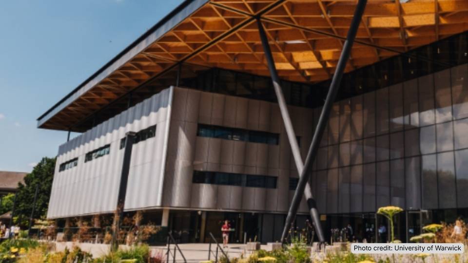 An exterior photo of a big modern building with a wooden roof on a sunny day with plants in the foreground.