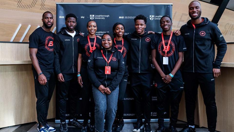 Group of people standing together in front of a banner with Loughborough University and Black in Sport Summit logos and wearing Black in Sport Summit merchandise.