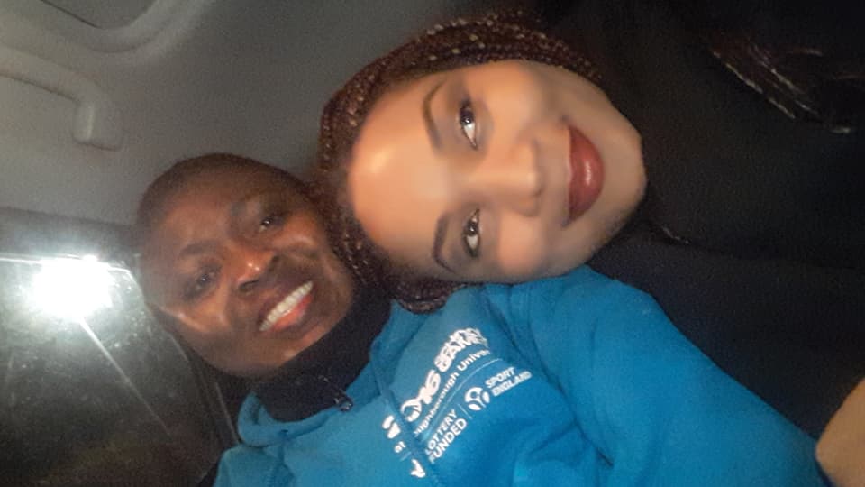 Close-up photo of two people sitting in a car and smiling, one is leaning their head on the other's shoulder.