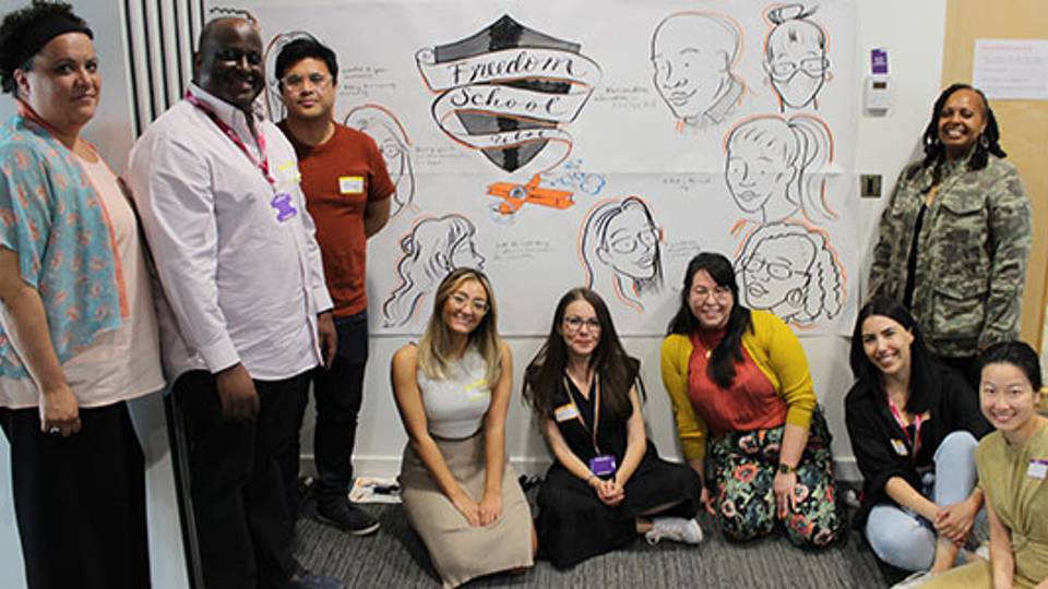 A group of people some standing and some sitting in front of a big poster with a 'Freedom School' badge on and drawings of faces.