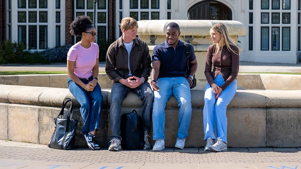 Four diverse university students sitting together on the edge of a large stone fountain on campus, talking and smiling on a sunny day.