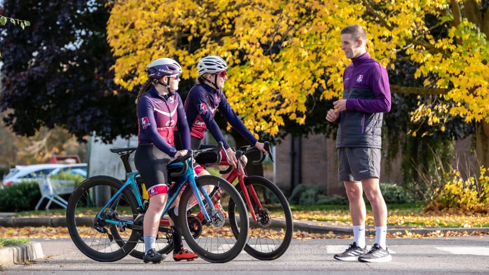 Two people on bikes talking to someone standing. In the background is a yellow autumnal tree.