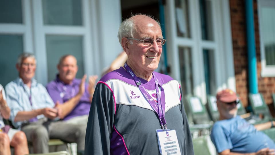 George Gandy wearing a Loughborough athletics tracksuit top. Standing in front of the athletics pavilion. There are people seated in the background. 