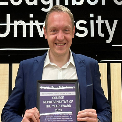 James wearing a suit and holding a certificate for a Course Representative of the Year Award. He stands in front of a Loughborough University logo on a wall with thin wooden panels.