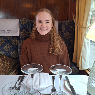 Lizzy sitting on a train smiling, a table is laid in front of her with wine glasses, plates and crockery on a white table cloth