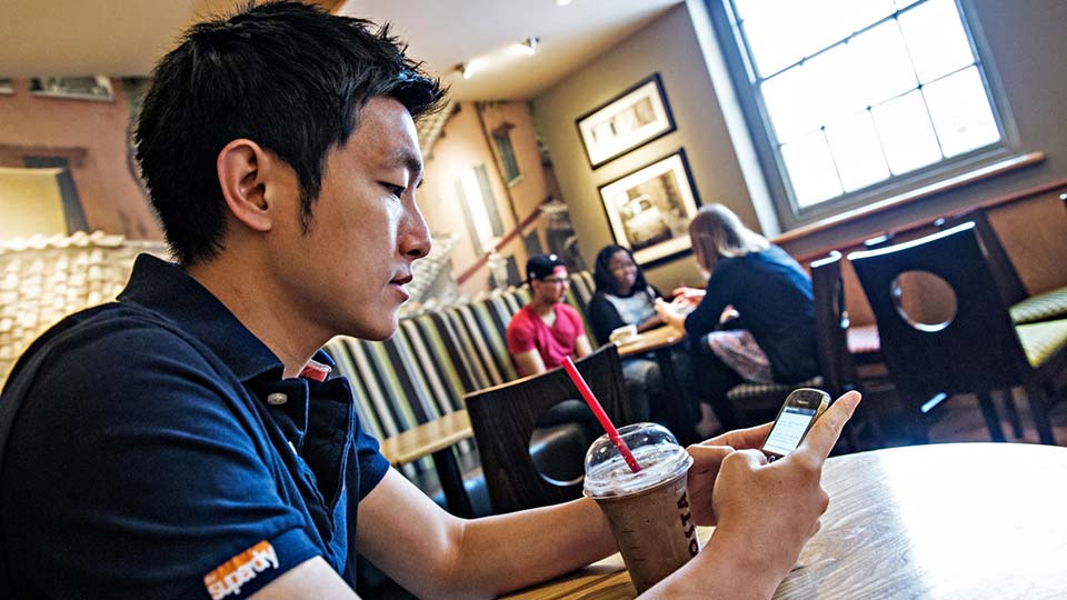 Student using a mobile phone while sitting in a café, with other students seated at tables in the background.