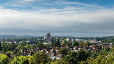 A leafy panorama view of Loughborough with houses, trees, and a few tall buildings under a partly cloudy sky, with hills visible in the background.
