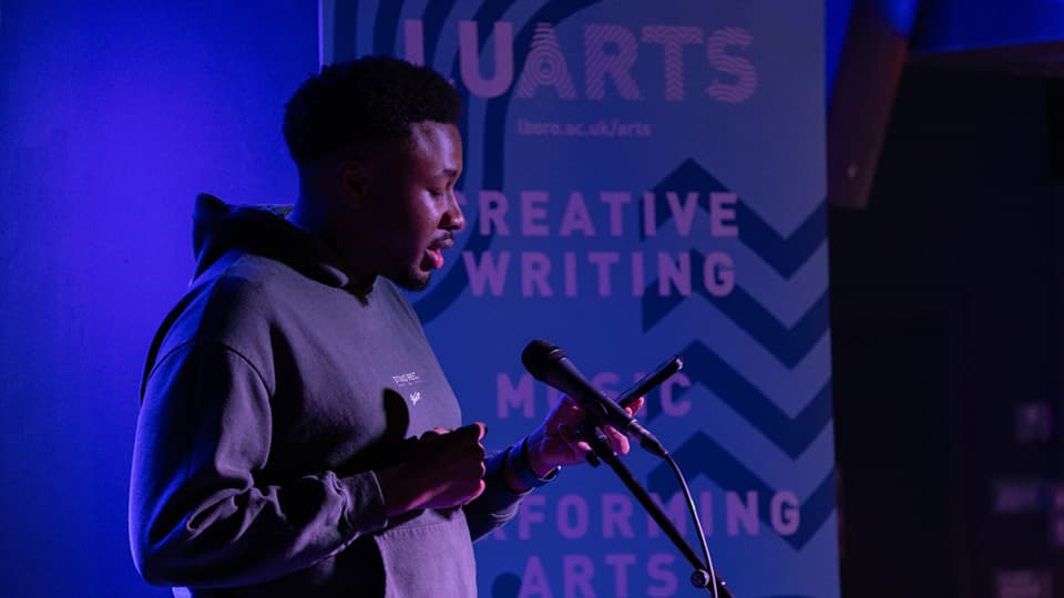 A man talking into a microphone, reading from his phone. There is purple lighting and he is wearing a grey hoodie.