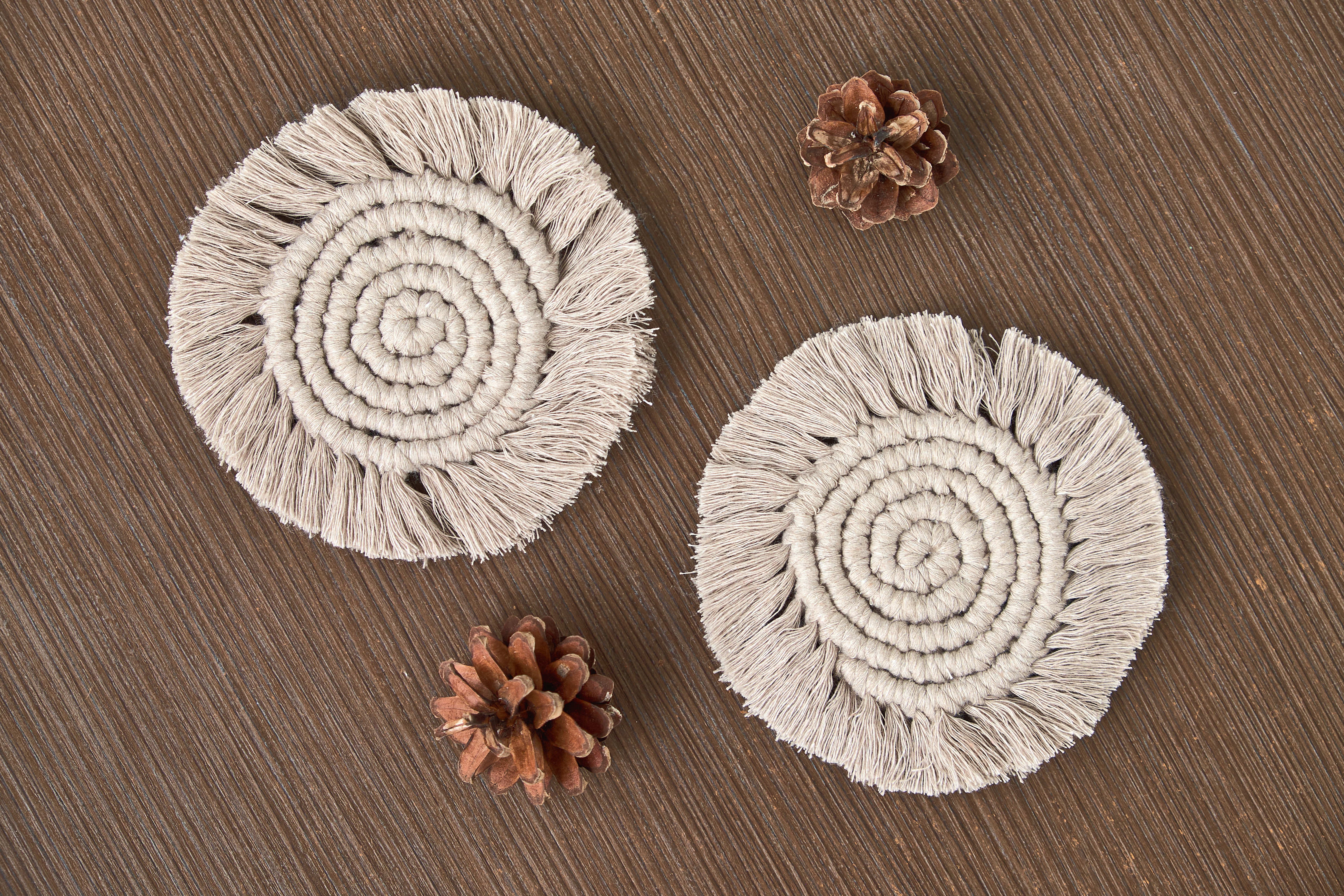 Two white macrame coasters on a wooden table, with two pine cones next to them. They are woven in a swirl shape, with tassel trims.