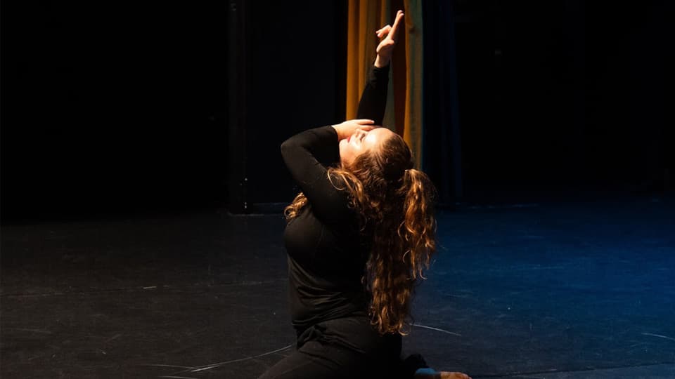 A dancer with long curly hair, wearing all black, kneeling on the floor with one arm pointed upwards.