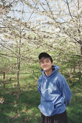 Student in early twenties in front of trees in blossom wearing a cap and hoodie and smiling at the camera with blue sky behind them