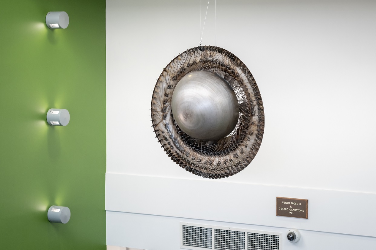 A hanging circular, planetary shaped welded steel sculputre. The walls in the background are green and white. There is a plaque on the wall behind the sculpture.