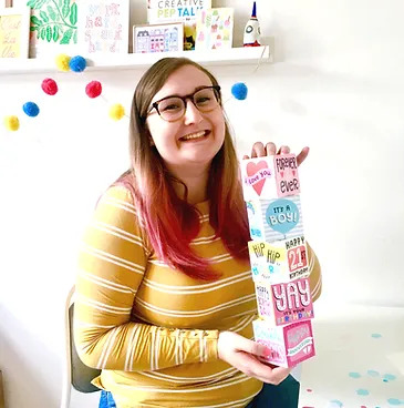 A smiling woman wearing glasses with long brown and red hair holding a stack of five illustrated boxes
