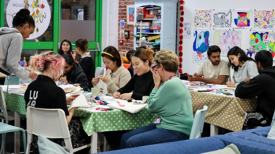 An arts workshop taking place with a mixed group of mostly female students sitting around tables that are covered in spotted tablecloths. They are drawing and stitching and the tables are covered in arts materials. One of the students is wearing a black polo shirt with 'LU Arts' on the back.