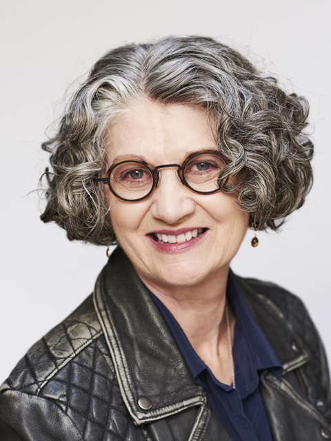 A middle aged female with short, curly dark grey hair and glasses, smiling