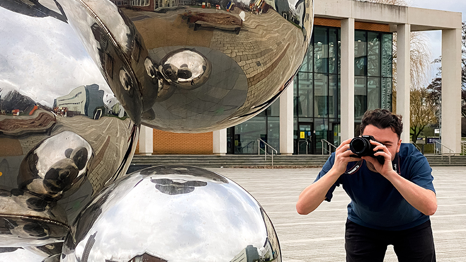 Close up of part of a stainless steel sculpture which has spheres stacked together with the surrounding buildings and features reflected in the steel. A white male with dark brown hair is at to the side of the sculpture taking a photo with a camera.