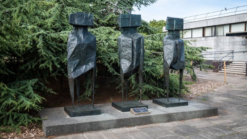 Bronze sculpture of 3 abstract figures, each with 3 legs, no arms and a rectangular 'heads'. They are pictured in front of a  cedar tree.