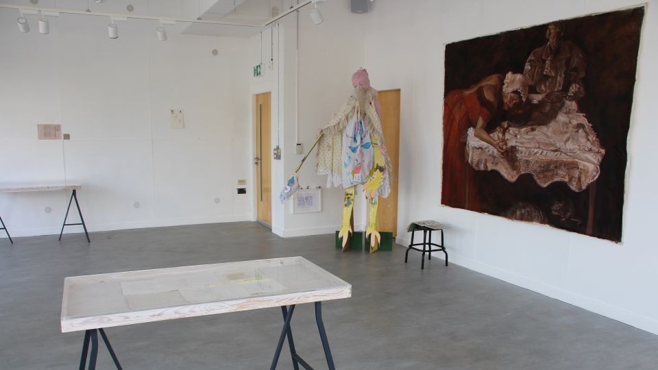 A gallery space with white walks and grey floor. A large unframed canvas painting is on the wall on the right with a birdlike textile sculptural piece hanging to the left of this and a display table in the foreground