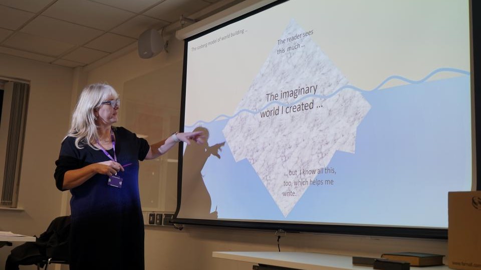 A female tutor with shoulder length hair and glasses is standing to the side of a smartboard and pointing to the screen