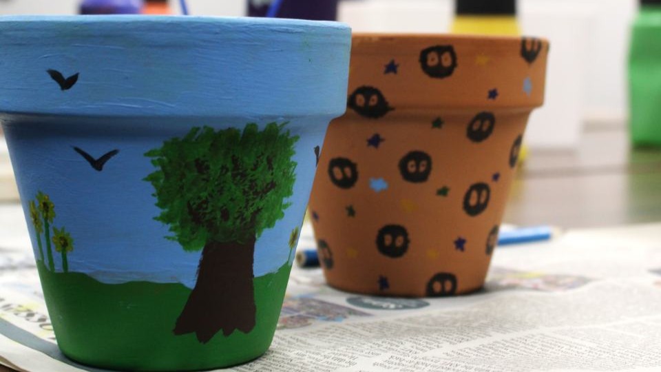 Two small terracotta flowerpots which have been painted. One has a large tree in leaf with blue sky and grass. The other is decorated with stars and small black characters that look like 'blobs' with large eyes