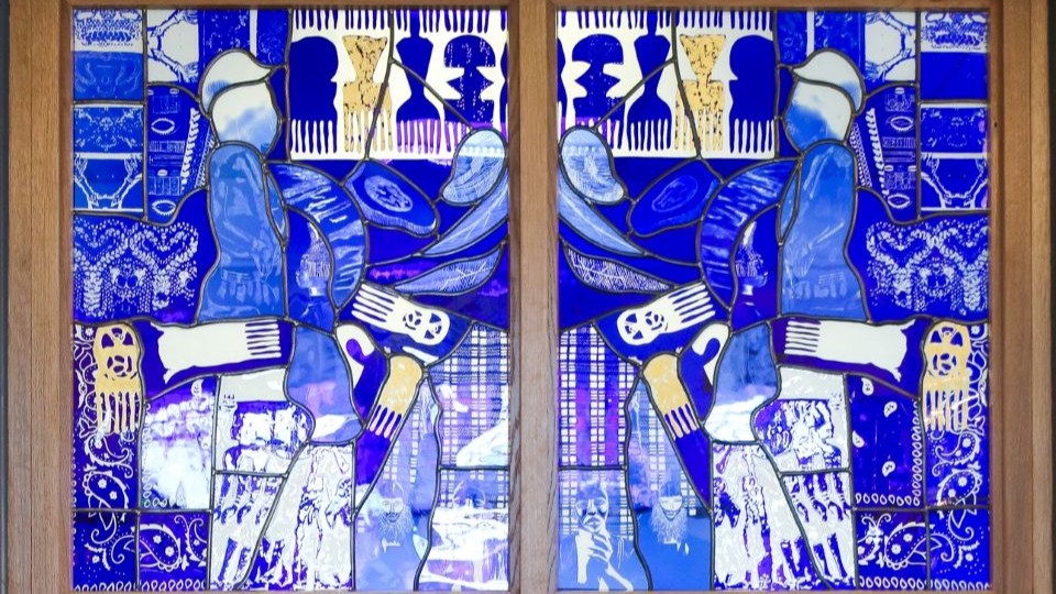 Stained glass window which is mostly blue with gold accents. The design is contemporary and includes different shapes of Afro hair combs and the outline of people as well as patterns
