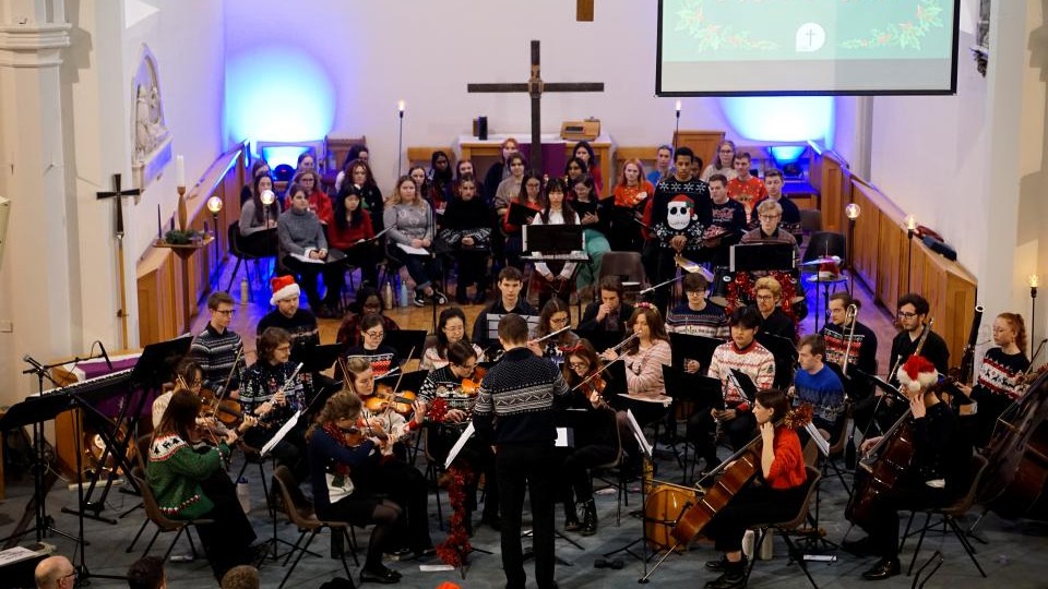A student symphony orchestra performing in a church wearing Christmas jumpers with a choir seated behind them