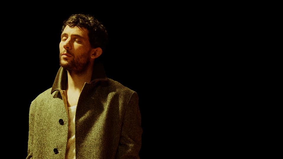 Dark background with a young white male standing to one side wearing a wool jacket with his eyes closed
