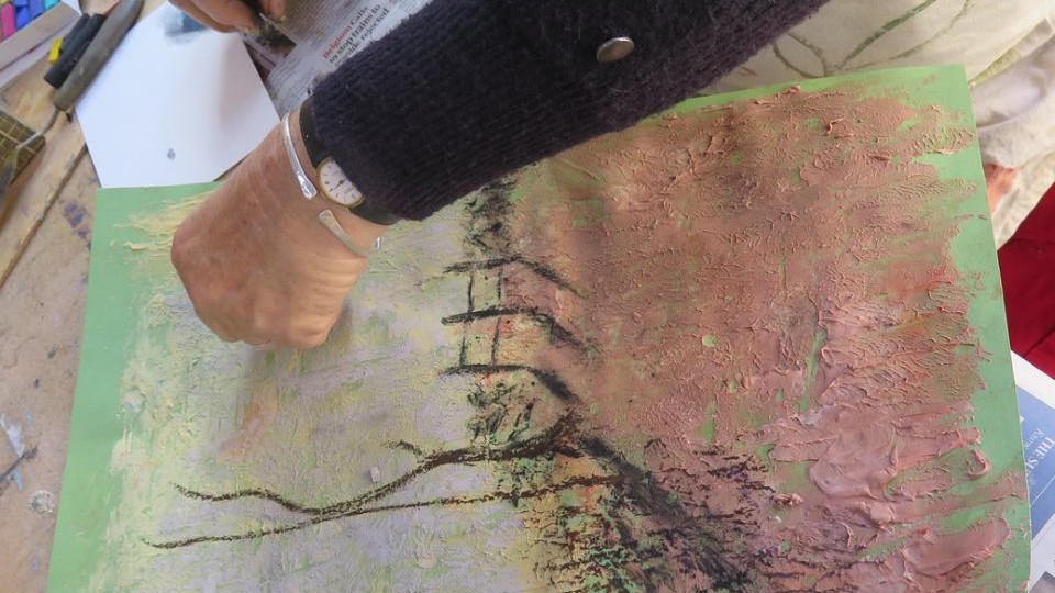 Close up of a landscape painting in progress with an arm across the top of the picture working on it. The paint is very textured and there is grey pastel/charcoal lines for a bare tree and a fence