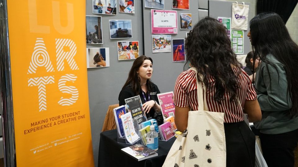 Bright orange LU Arts banner to the side of a table displaying various poster and leaflet holders. A female student is sat behind the table talking to people with photos on a displayboard behind her
