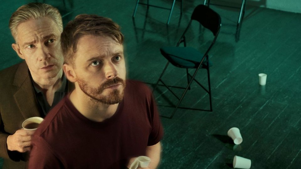 Two male actors - one in their late 20s/early 30s and the other in their 40s/50s standing one behind the other looking upwards. They are in an empty room with chairs and paper cups lying on the floor.