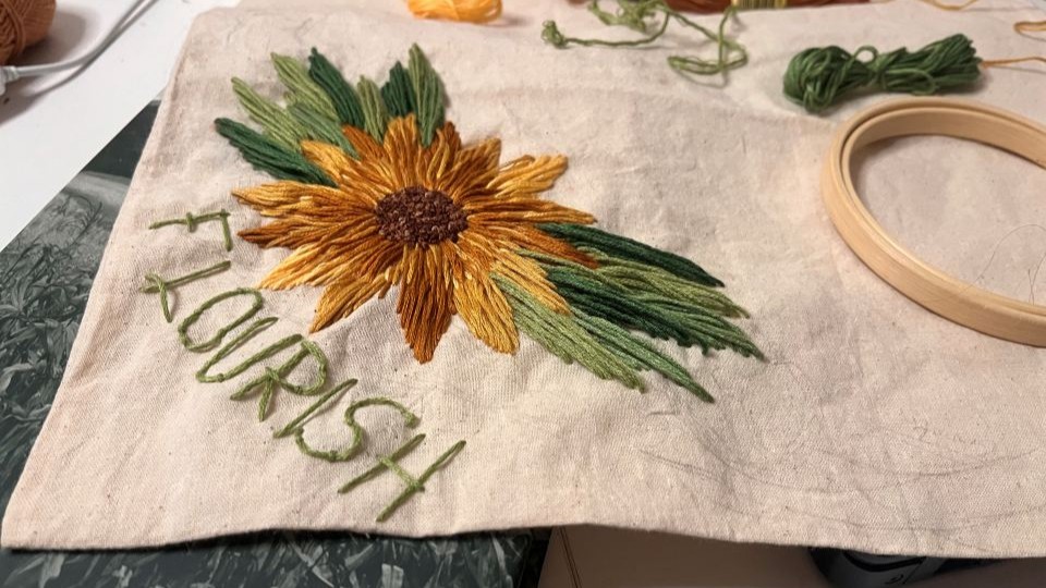 Close up of an embroidered sunflower on plain calico fabric with the word 'Flourish' stitched beneath it