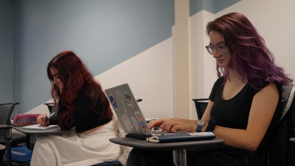 A writing class in progress with two female students seated on chairs with arm tables. One is writing in a book and the other is typing on a laptop