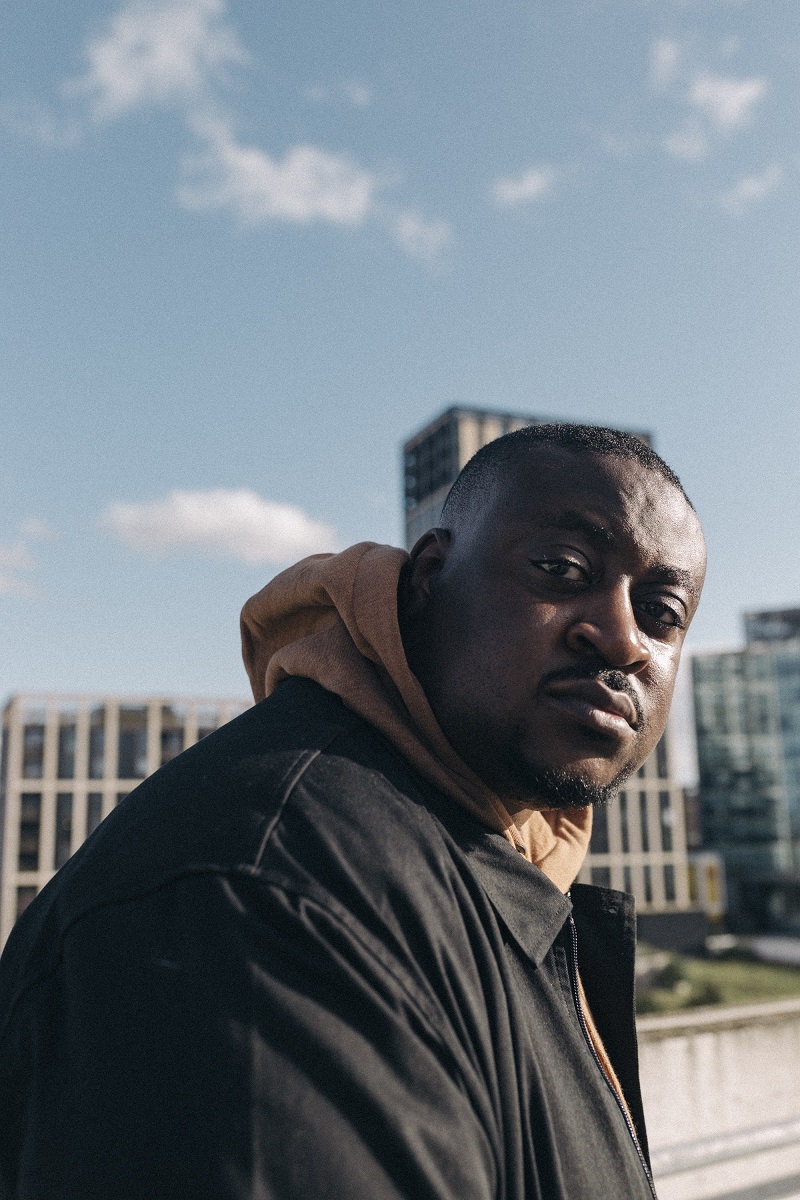 A photograph of a black man in his mid 30s against a blue sky. He is looking at the camera.