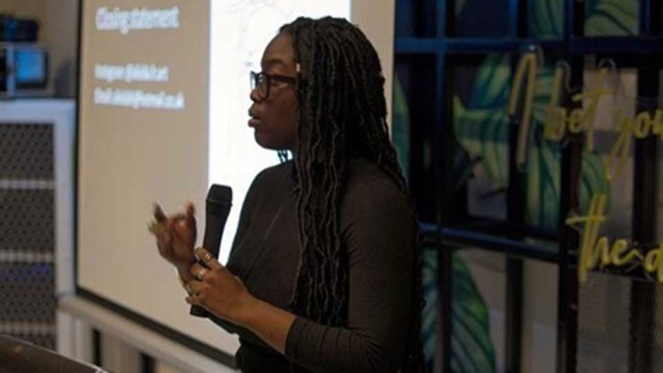Young female with dark brown skin and long black braided hair wearing glasses and holding a microphone in front of a projector screen. 