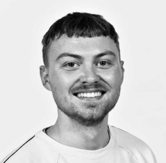 Head shot of white young male looking directly at camera and with short brown hair and a light moustache/beard, photographed in black and white.