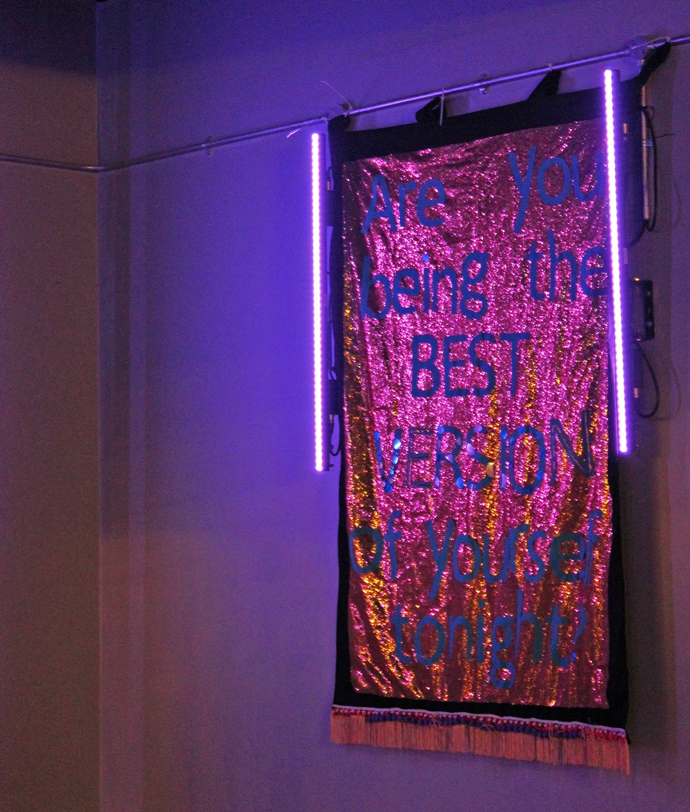 A sequined banner on a wall in a purple lit club room saying Are You Being The Best Version Of Yourself Tonight