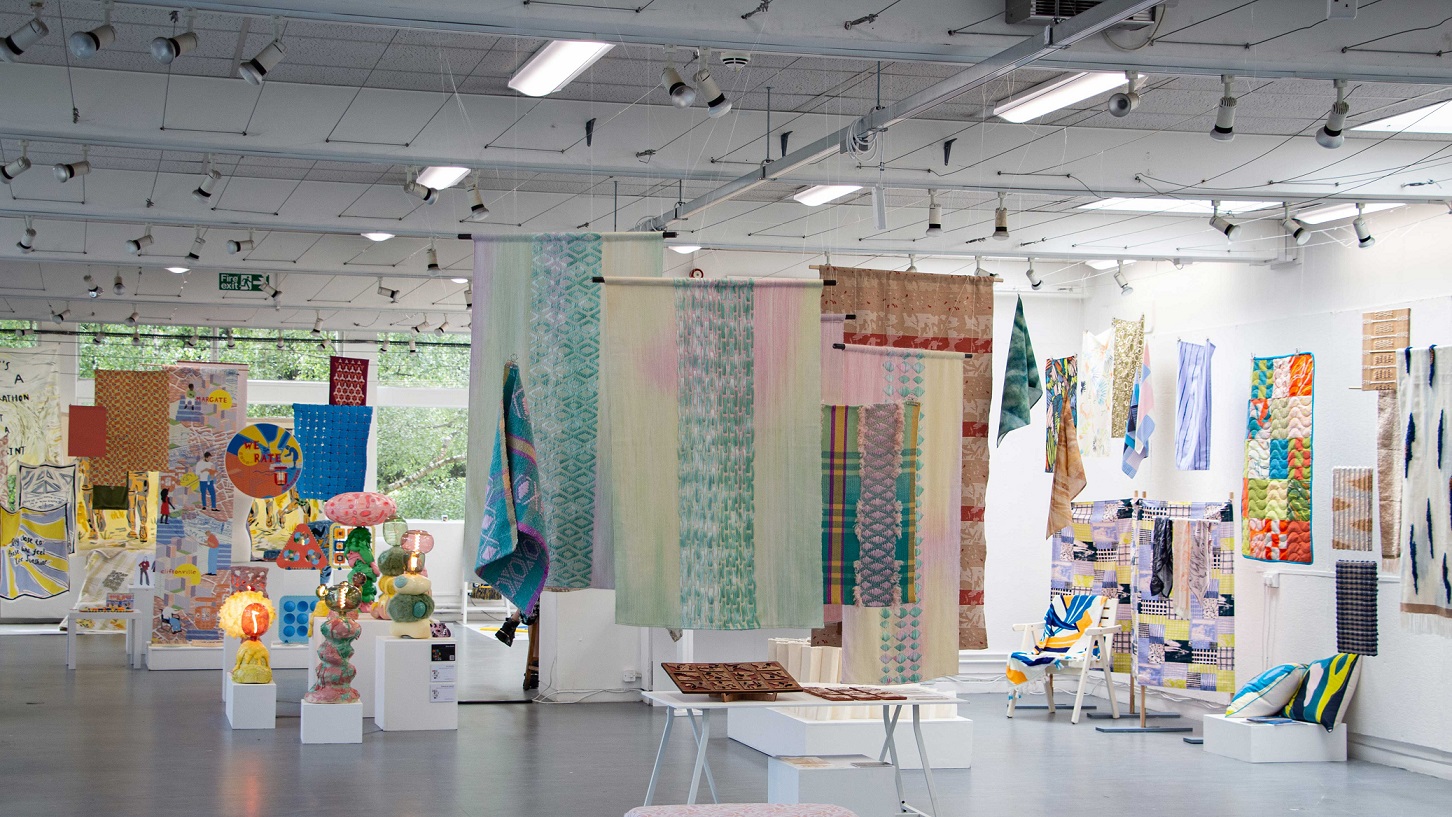 Work by various textiles students is hanging and displayed on plinths in the Arts Degree Show. It is a bright and airy room with one white wall and one window wall. Woven textile work in mainly pastel colours are hanging from the ceiling and on walls. There is a collection of brightly coloured ceramic lamps made up of different spherical and abstract shaped pieces stacked on top of one another.