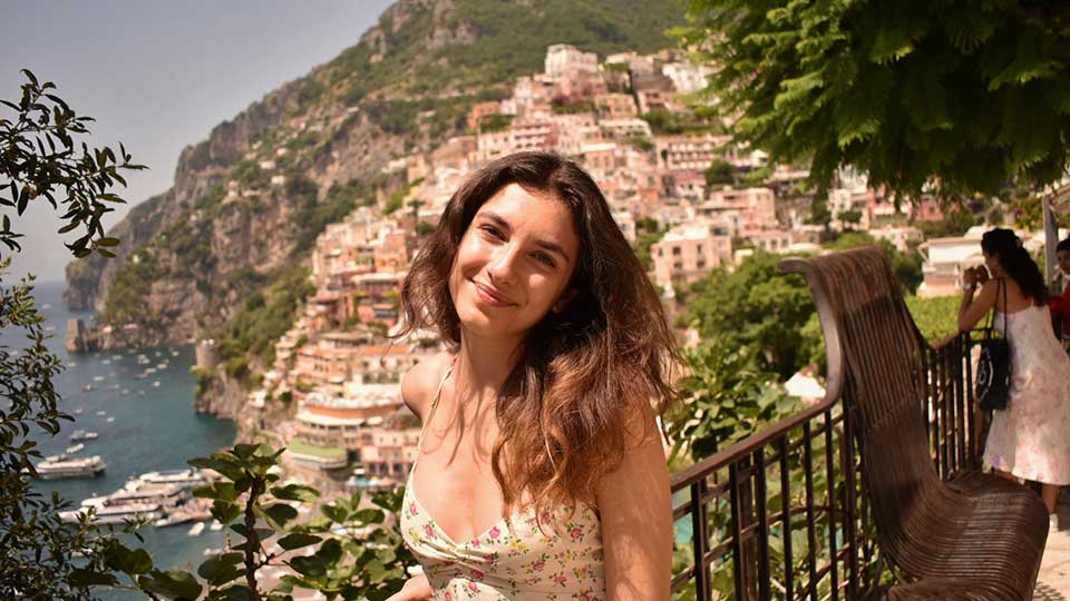 Young white female with long brown hair, wearing a sundress and smiling at the camera. She is pictured on a hill side overlooking a hillside coastal town somewhere in the Mediterranean.