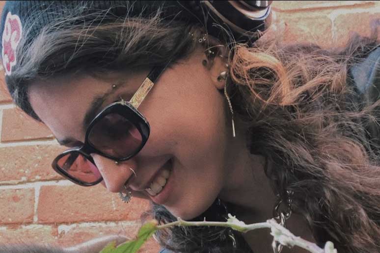 Headshot taken at a diagonal angle of a young female looking down and away from the camera. She is smiling, wearing a black knitted hat, over the ear headphones (just visible), black sunglasses and has long wavy brown hair. She has multiple nose and ear piercings with a stud and rings.